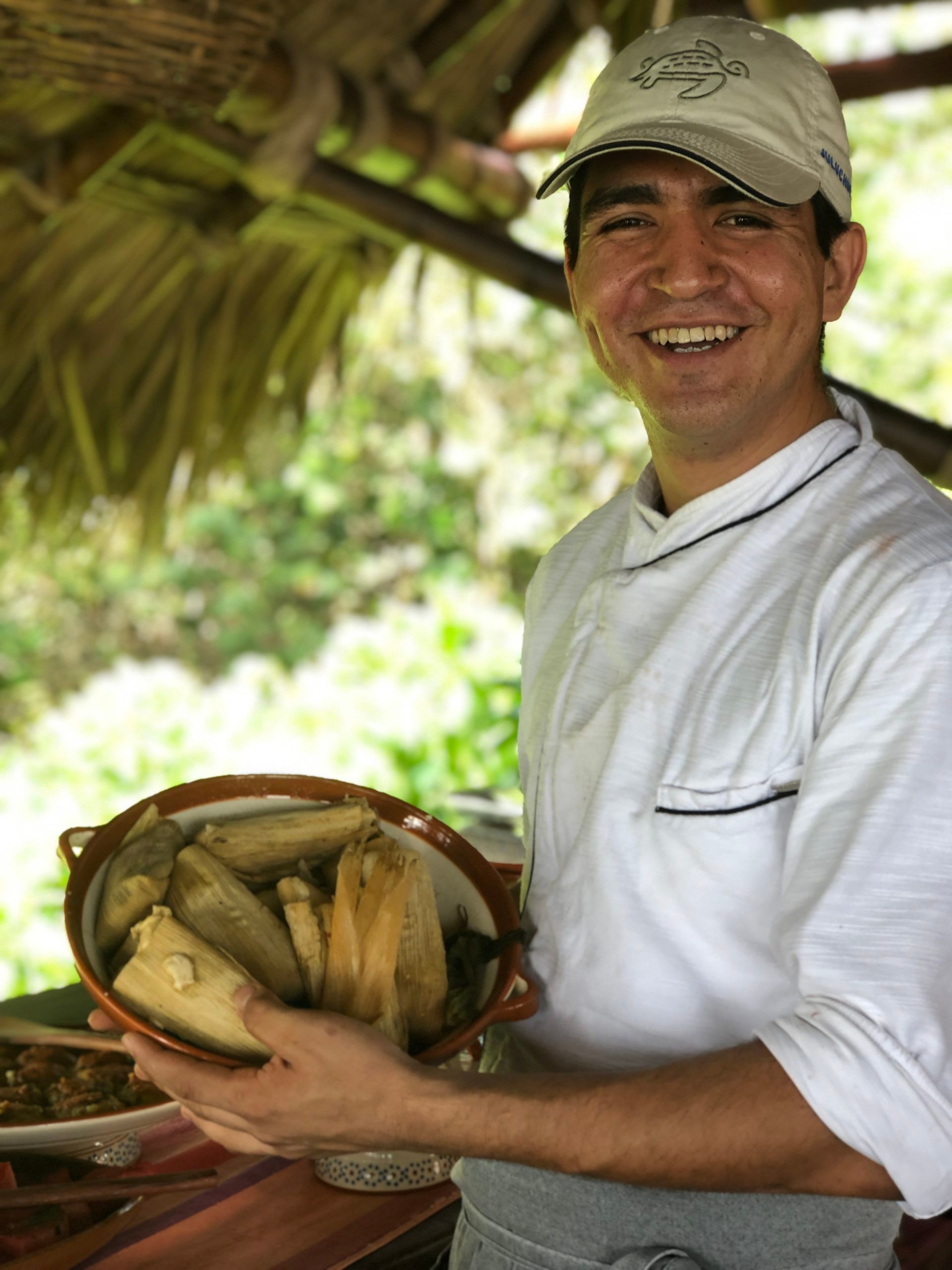 Smiling man in a white shirt holding a basket of bread or produce under a thatched roof