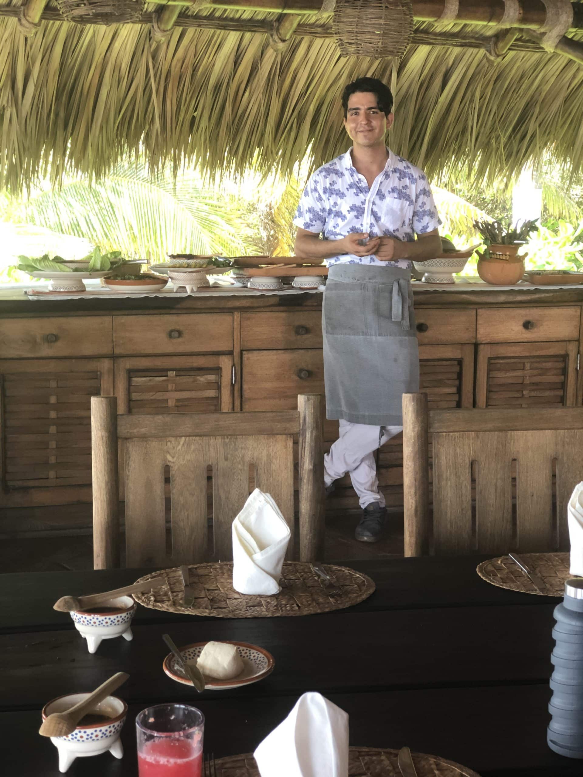 Server behind a rustic bar in a thatched-roof restaurant, with tables set in the foreground.