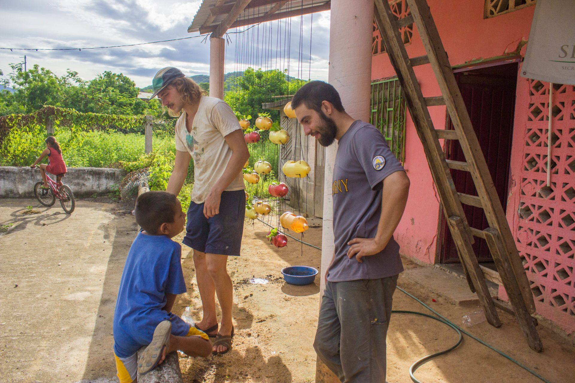 Three people standing outside a rustic house, one kneeling and another talking to a child in a blue shirt.
