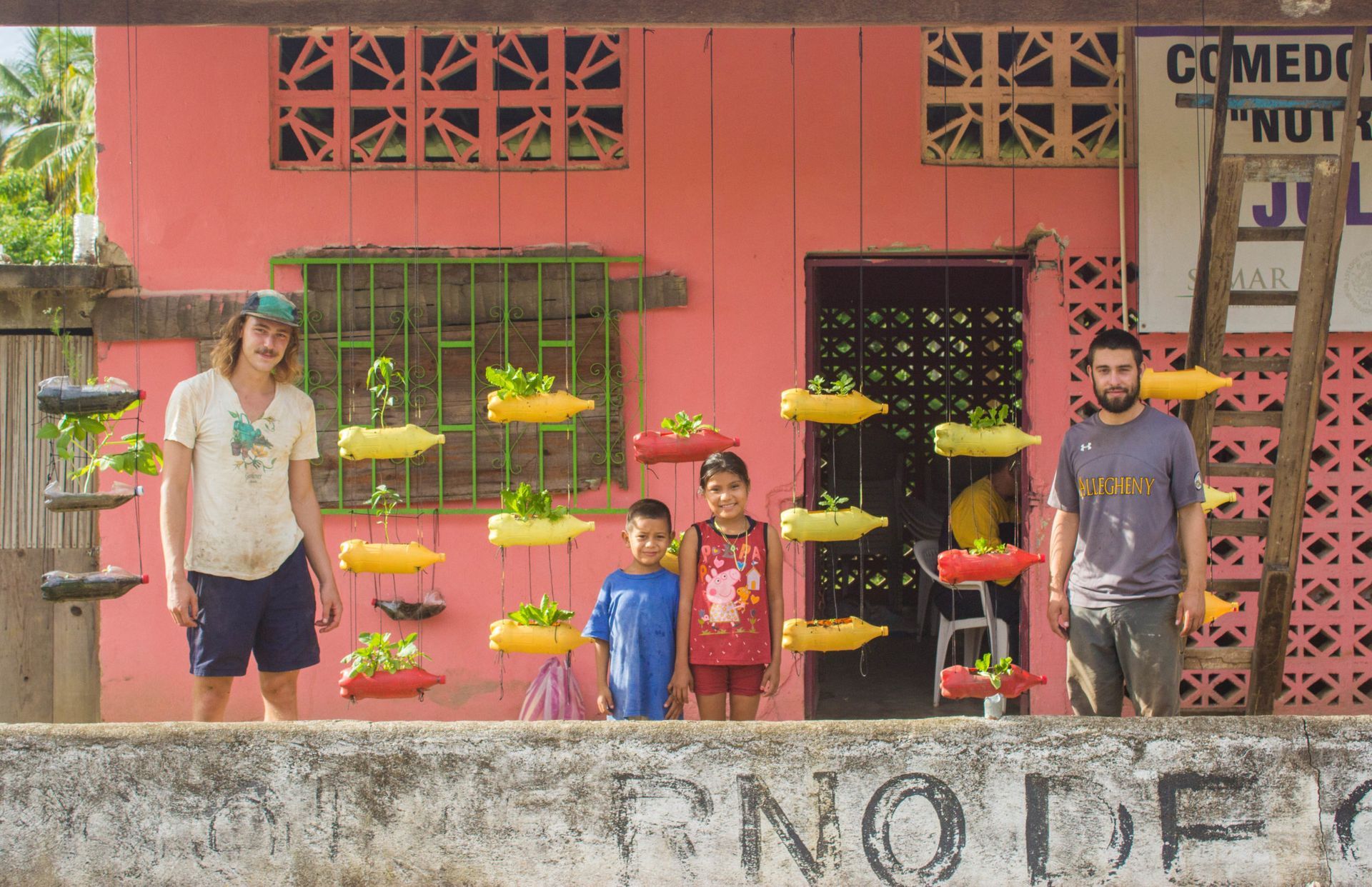 Four people standing by a pink wall with hanging yellow flower pots in a garden area