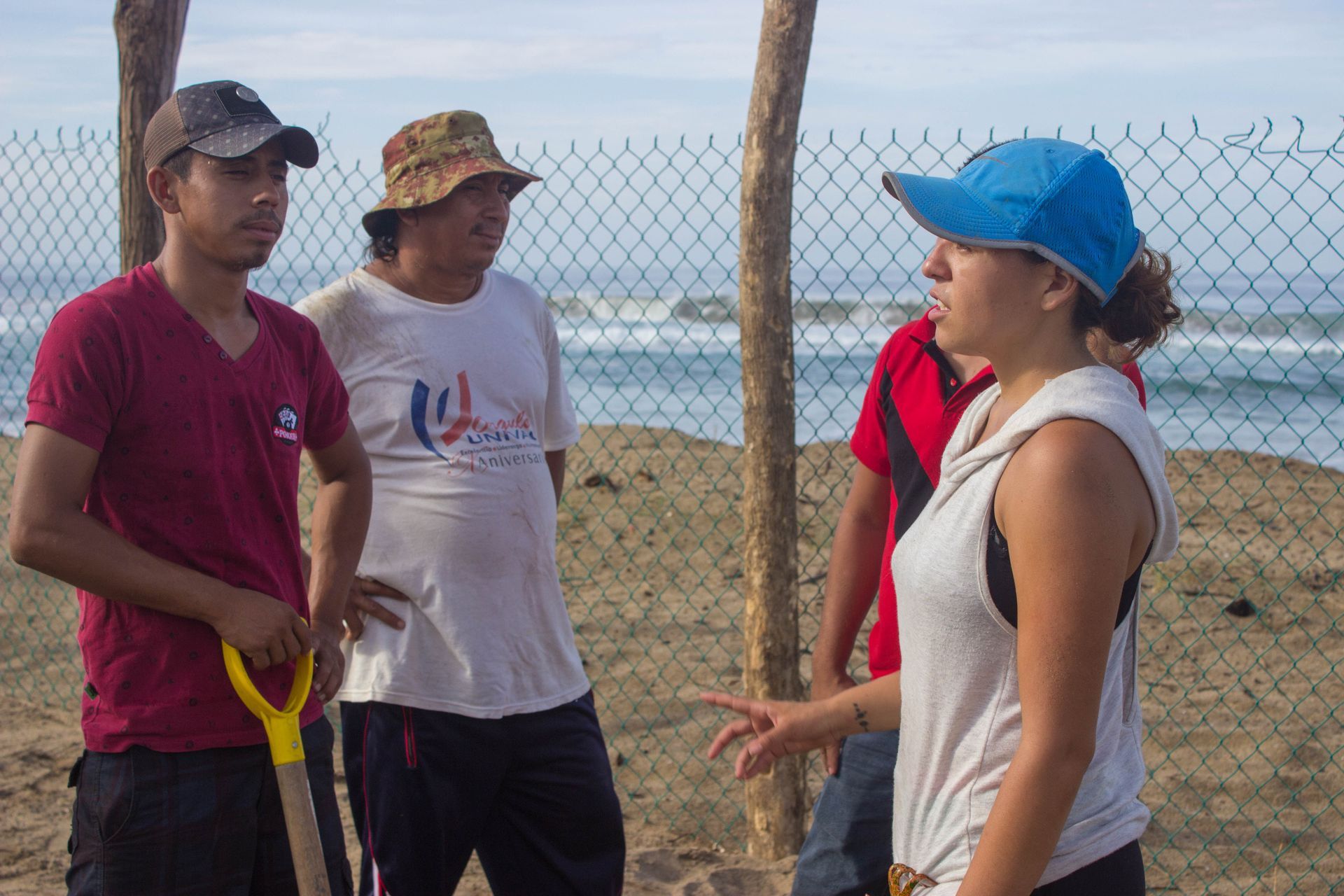 Three people talking outdoors by a chain-link fence near the beach