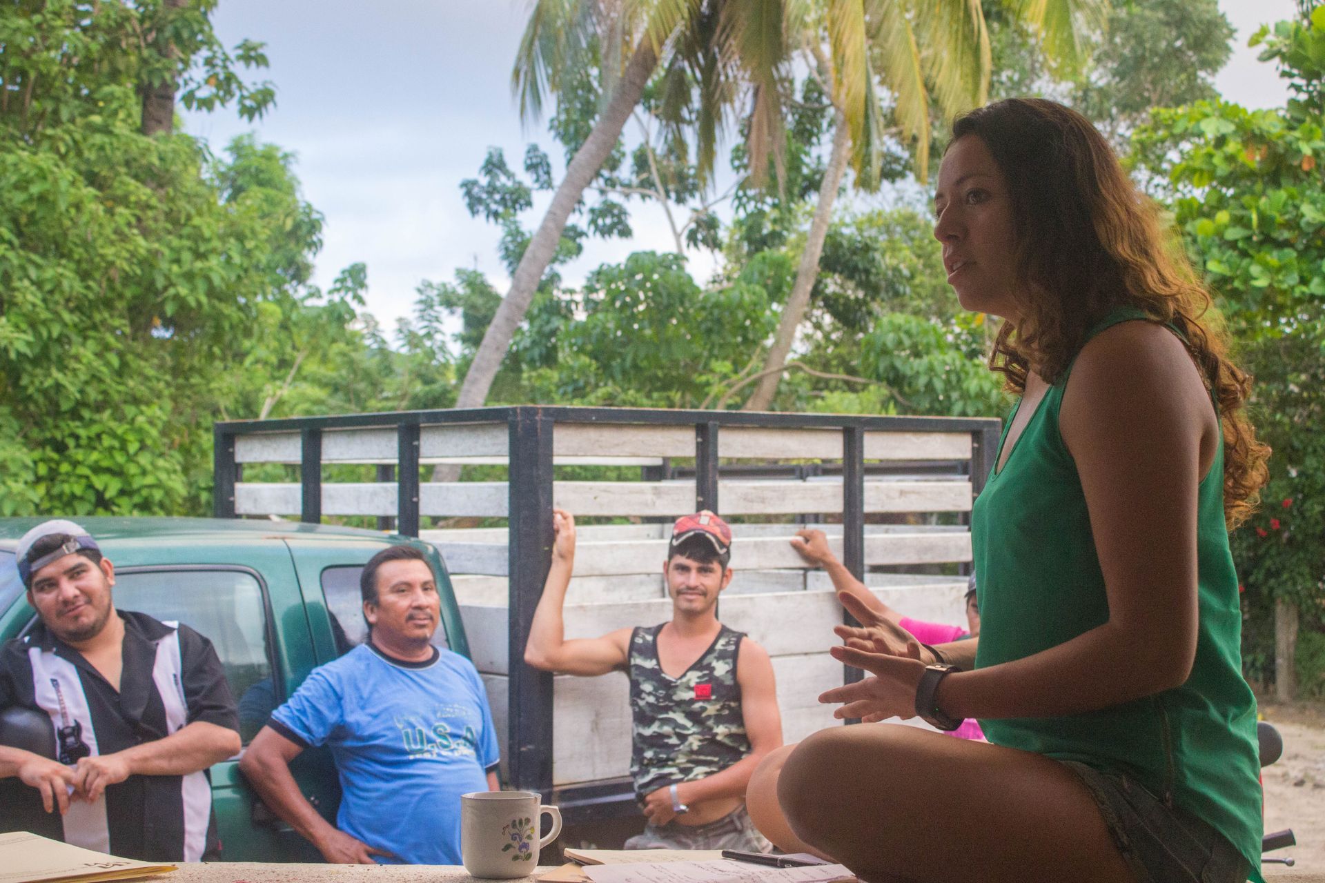 Group sitting outdoors on a patio, chatting beside a woman in a green dress under palm trees.