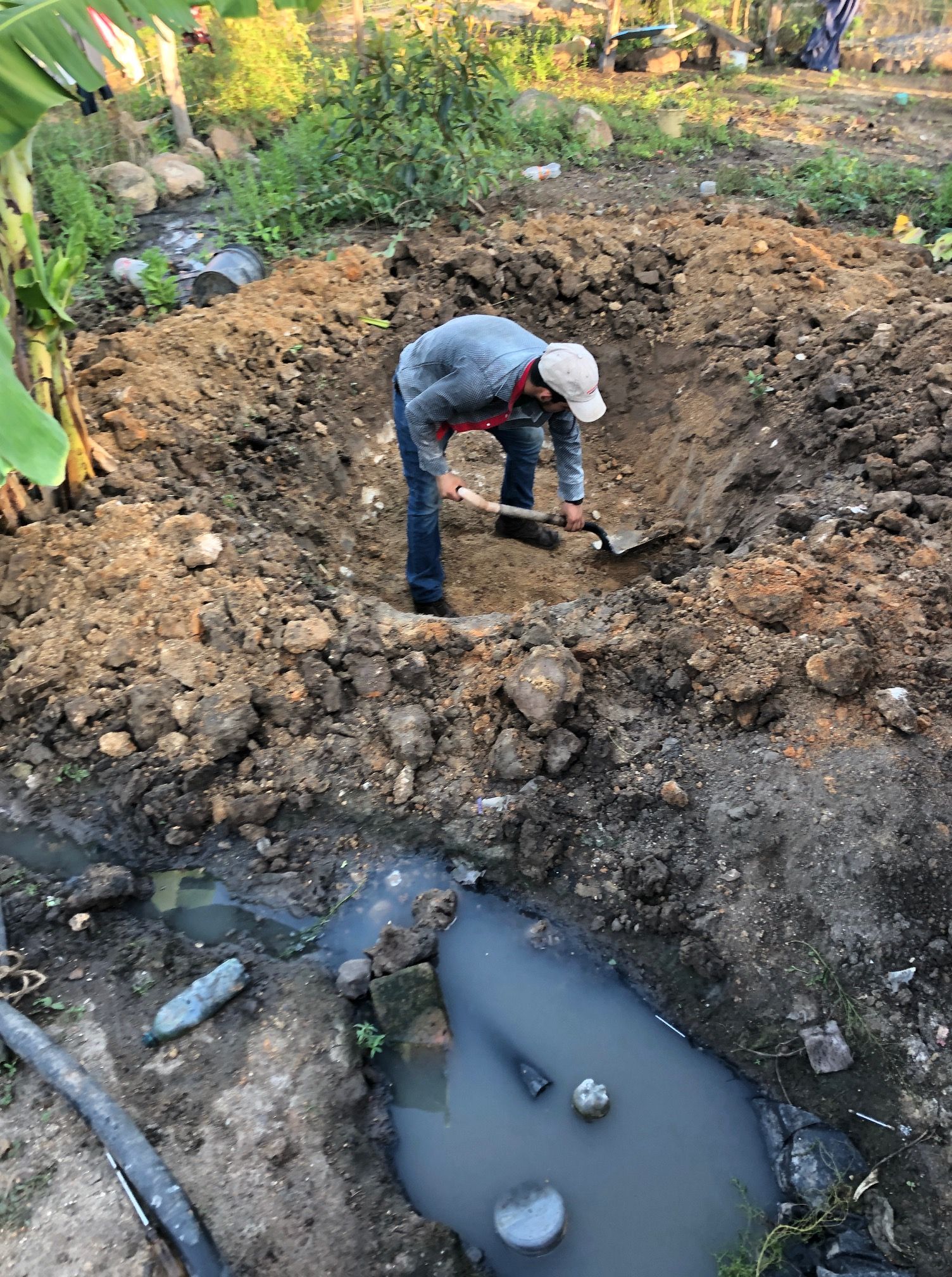 Worker digging a large muddy hole beside dark water in an outdoor construction site