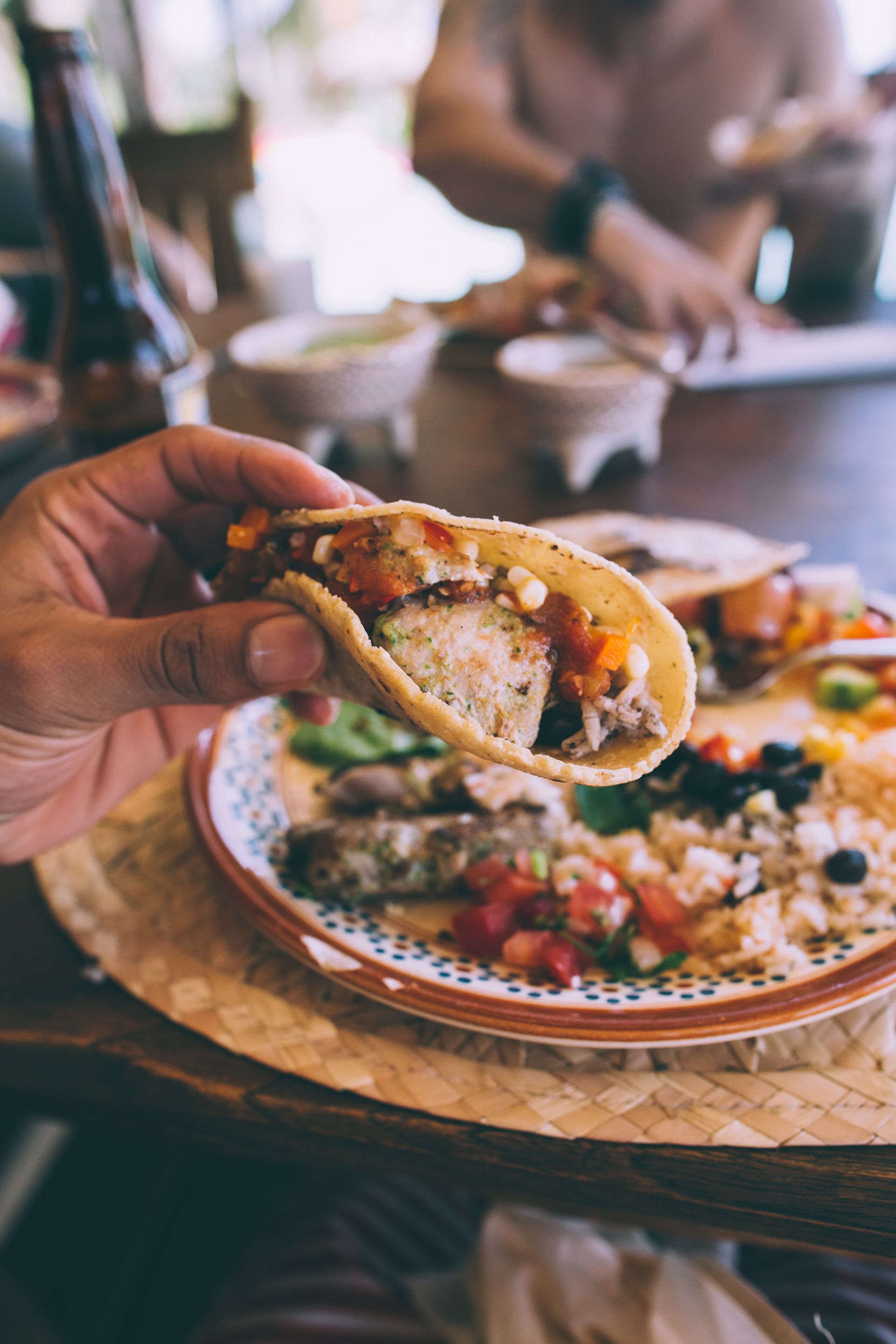 Hand holding a taco over a plate of assorted tacos on a restaurant table