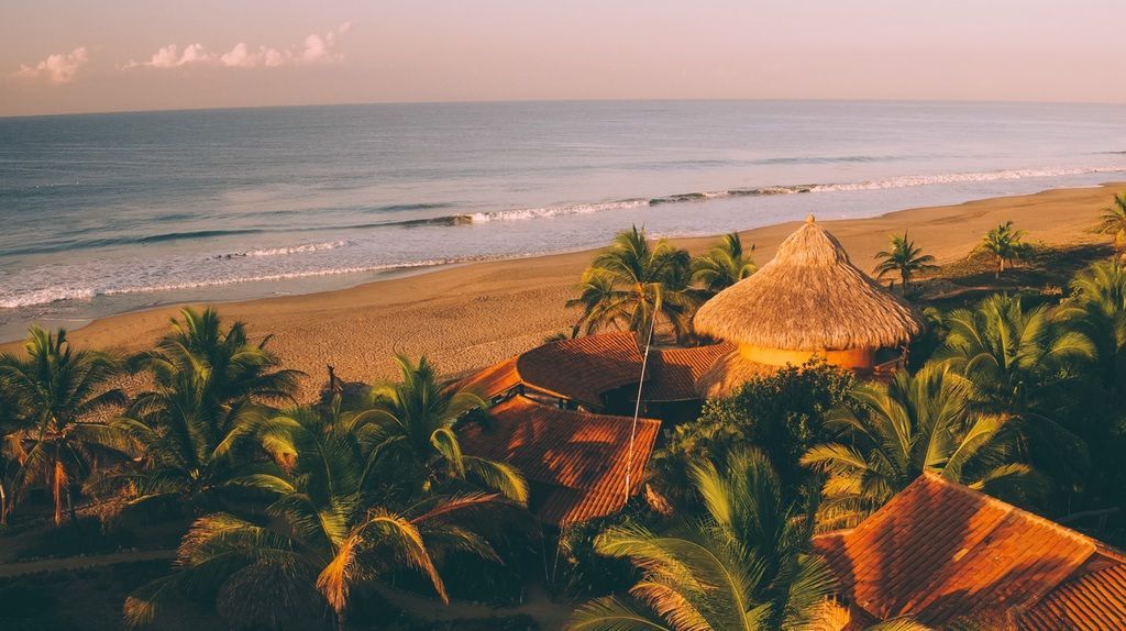 Tropical beach resort with palm trees, thatched roofs, and calm ocean at sunset