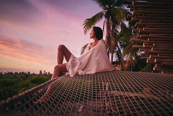 A woman is sitting in a hammock looking at the sunset.