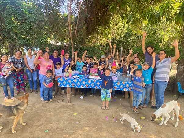 Group gathered outdoors around a blue table, smiling and raising hands in a leafy yard.