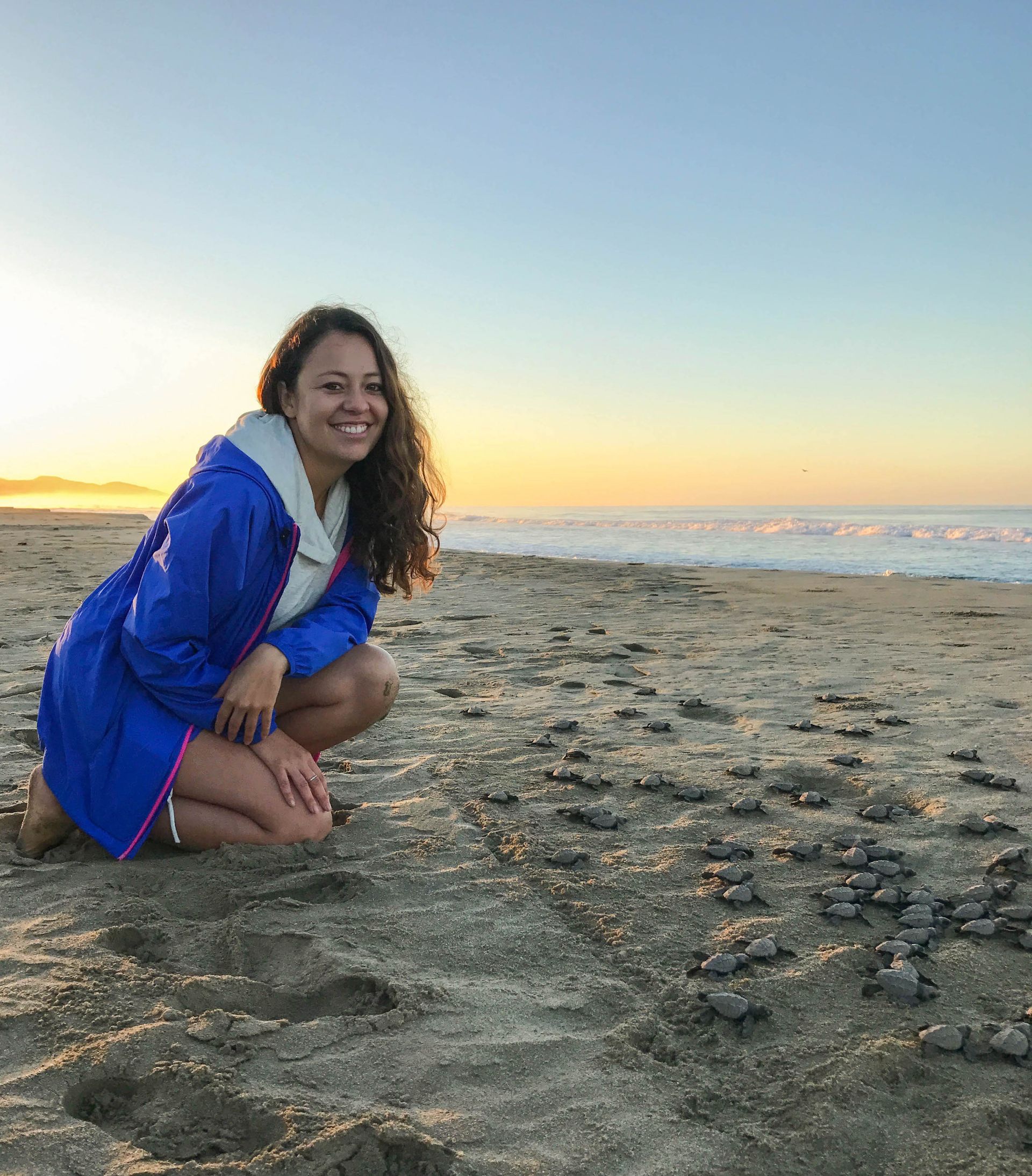 Woman crouching on a sandy beach at sunset, smiling in a blue jacket near the ocean.