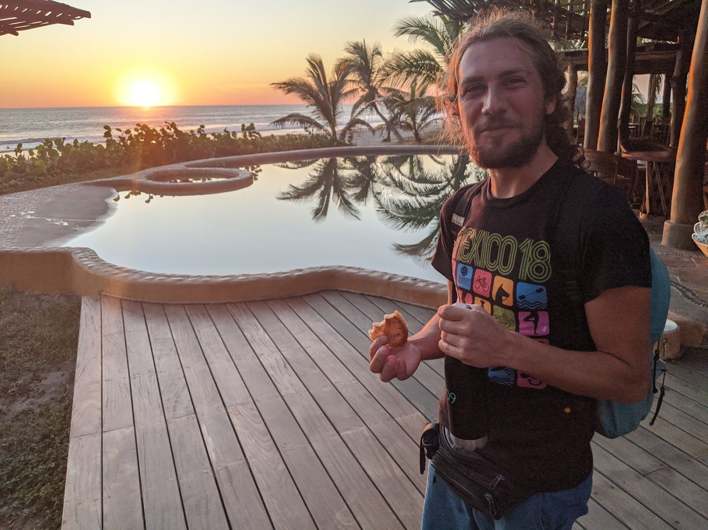 Man standing by a resort pool at sunset, holding food with palm trees and ocean in the background.