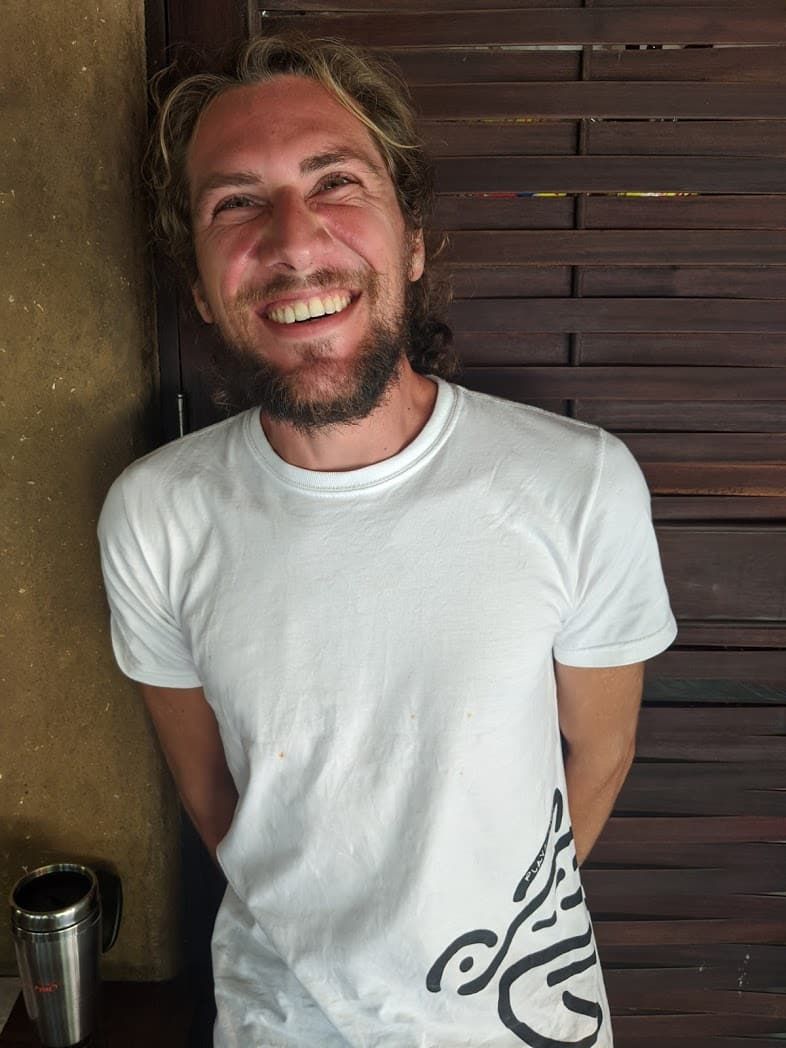 Smiling person in a white T-shirt standing indoors against a wooden wall.