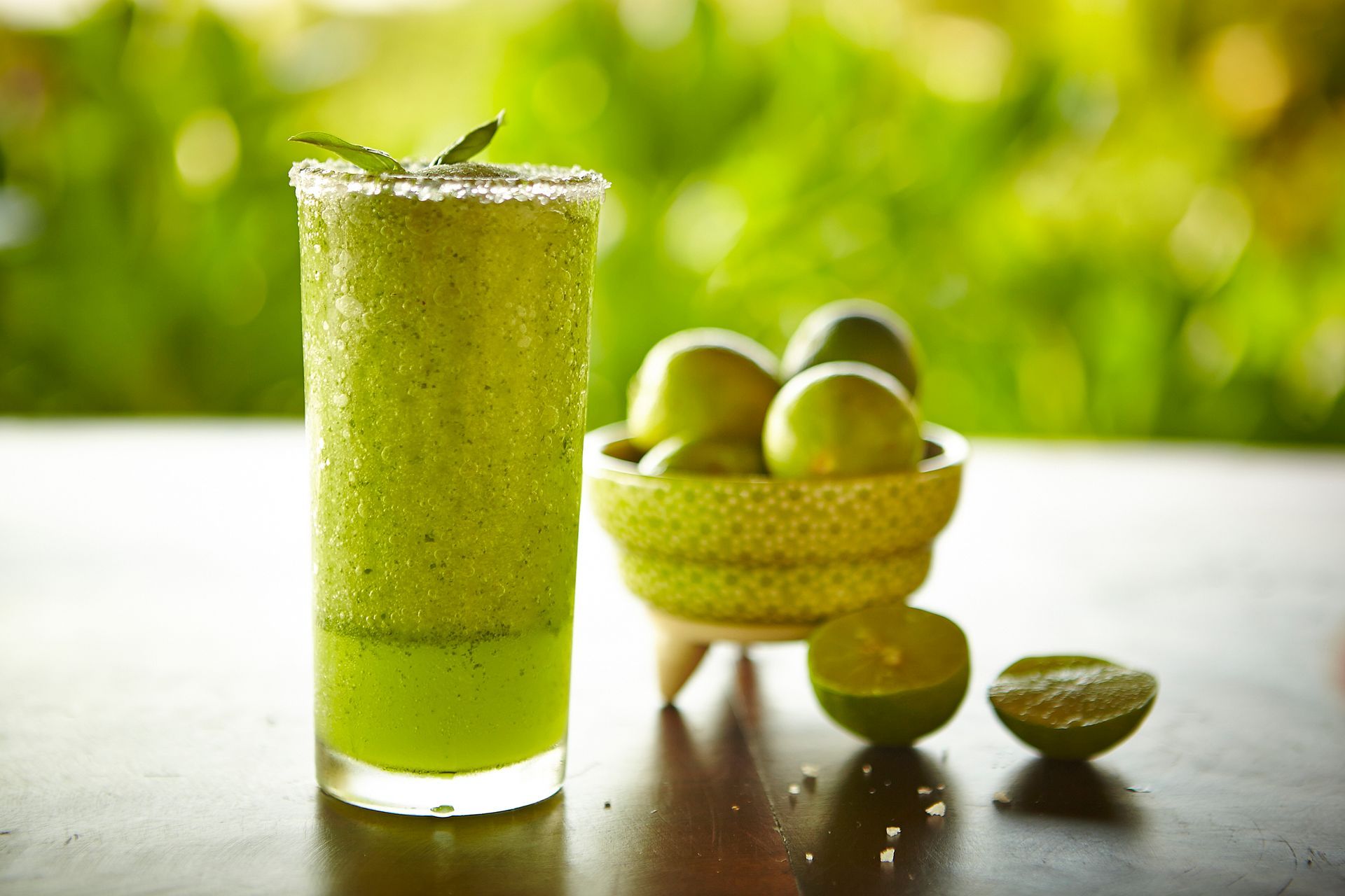 Green smoothie in a tall glass beside a bowl of limes on a sunlit table
