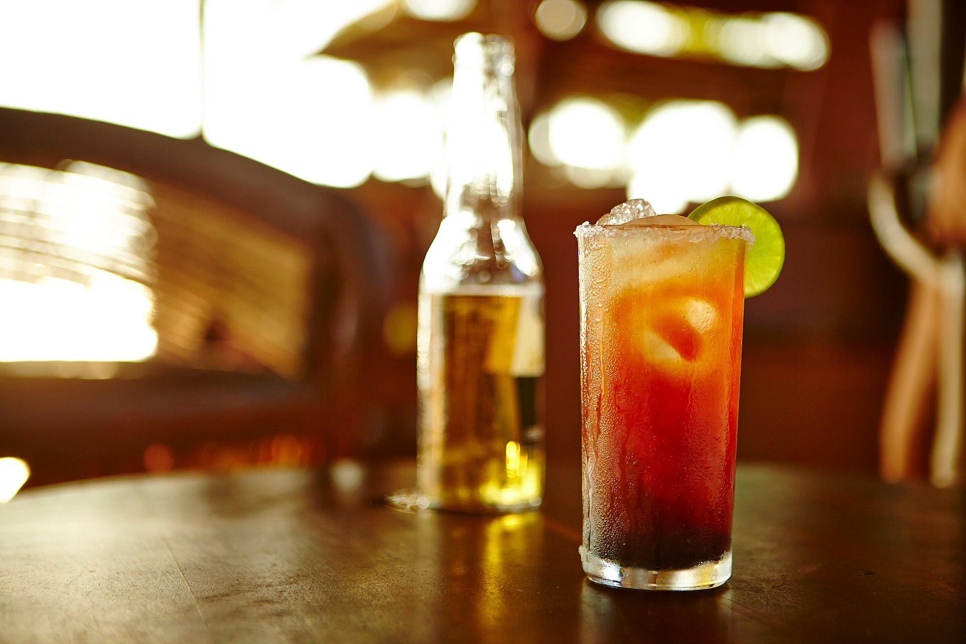 Glass of red cocktail with lime on a bar table, with a beer bottle blurred in the background