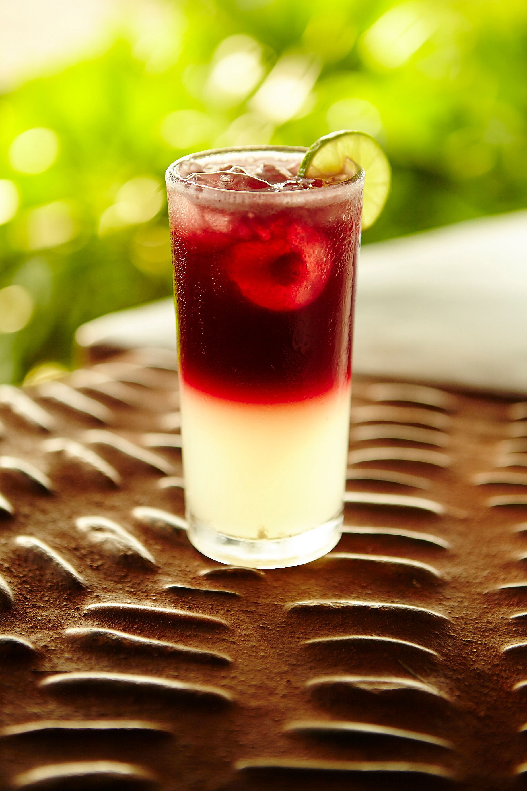 Tall red and white layered drink with a lime slice on a woven table outdoors