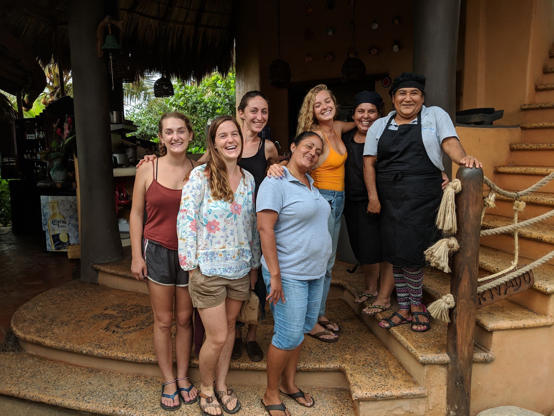 Six smiling people standing together on a porch with steps and a rope railing.