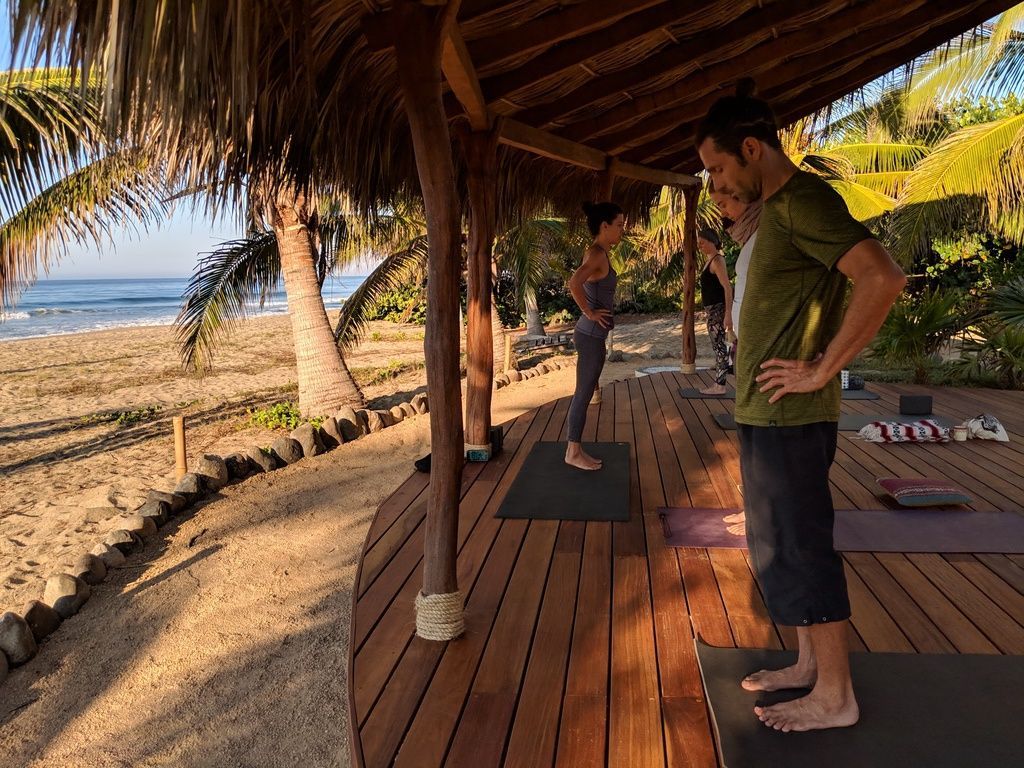 People practicing yoga on a wooden beach deck under a thatched roof by the ocean.