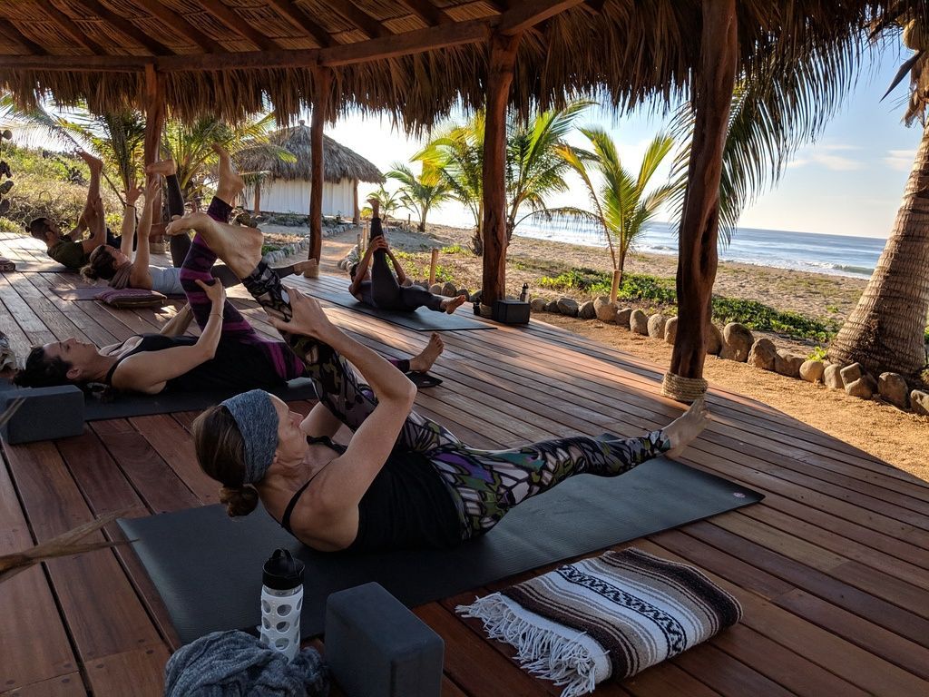 People doing yoga on mats under a thatched seaside cabana with palm trees and ocean views