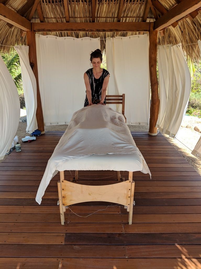 Massage table under a thatched gazebo with sheer curtains, overlooking a wooden deck.