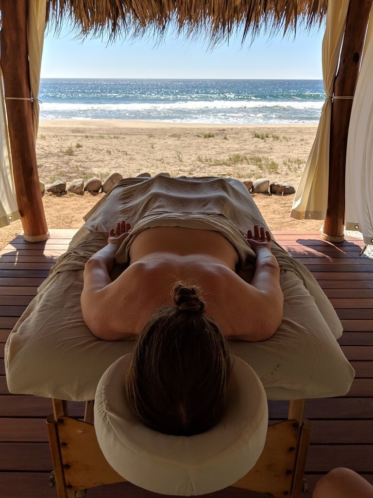 Person lying on a massage table under a thatched canopy, facing a sunny beach and ocean.