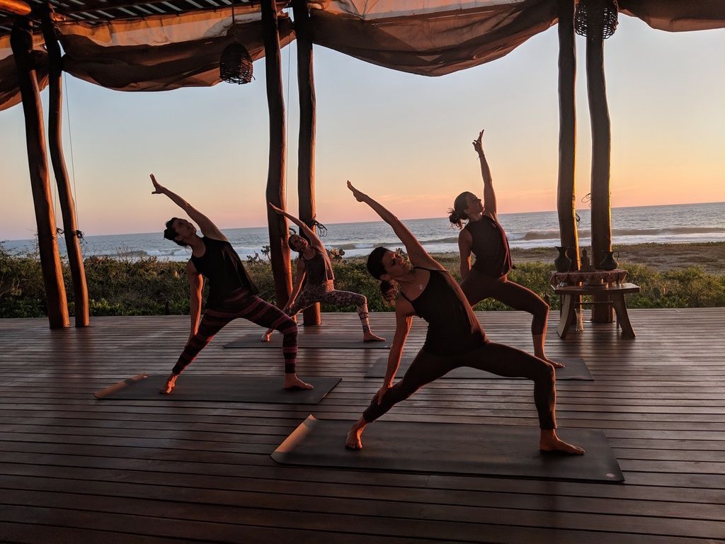 People doing yoga poses on a wooden deck at sunset, with ocean in the background.