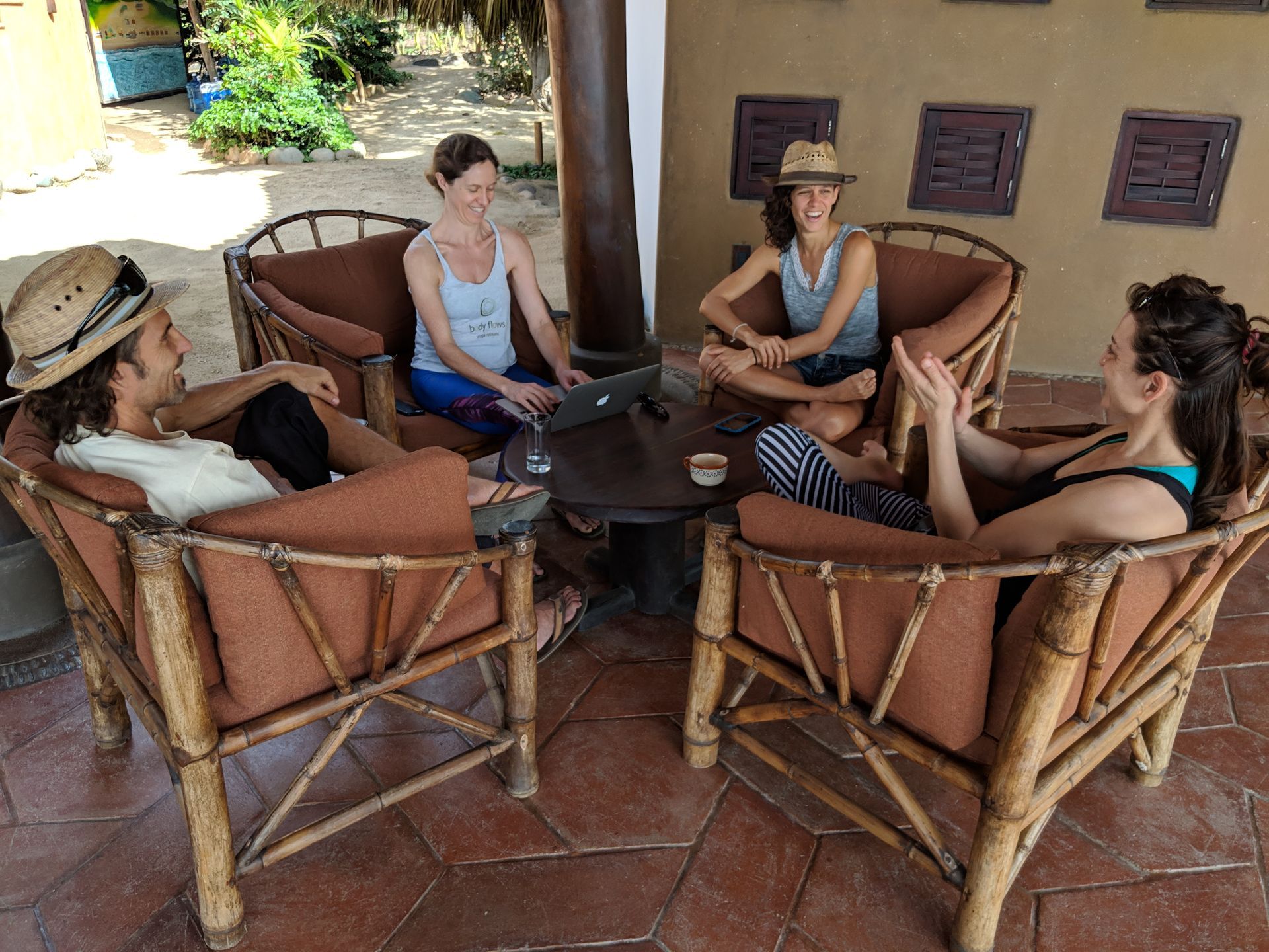 Four people sit in wicker chairs around a table on a covered patio, chatting and relaxing.