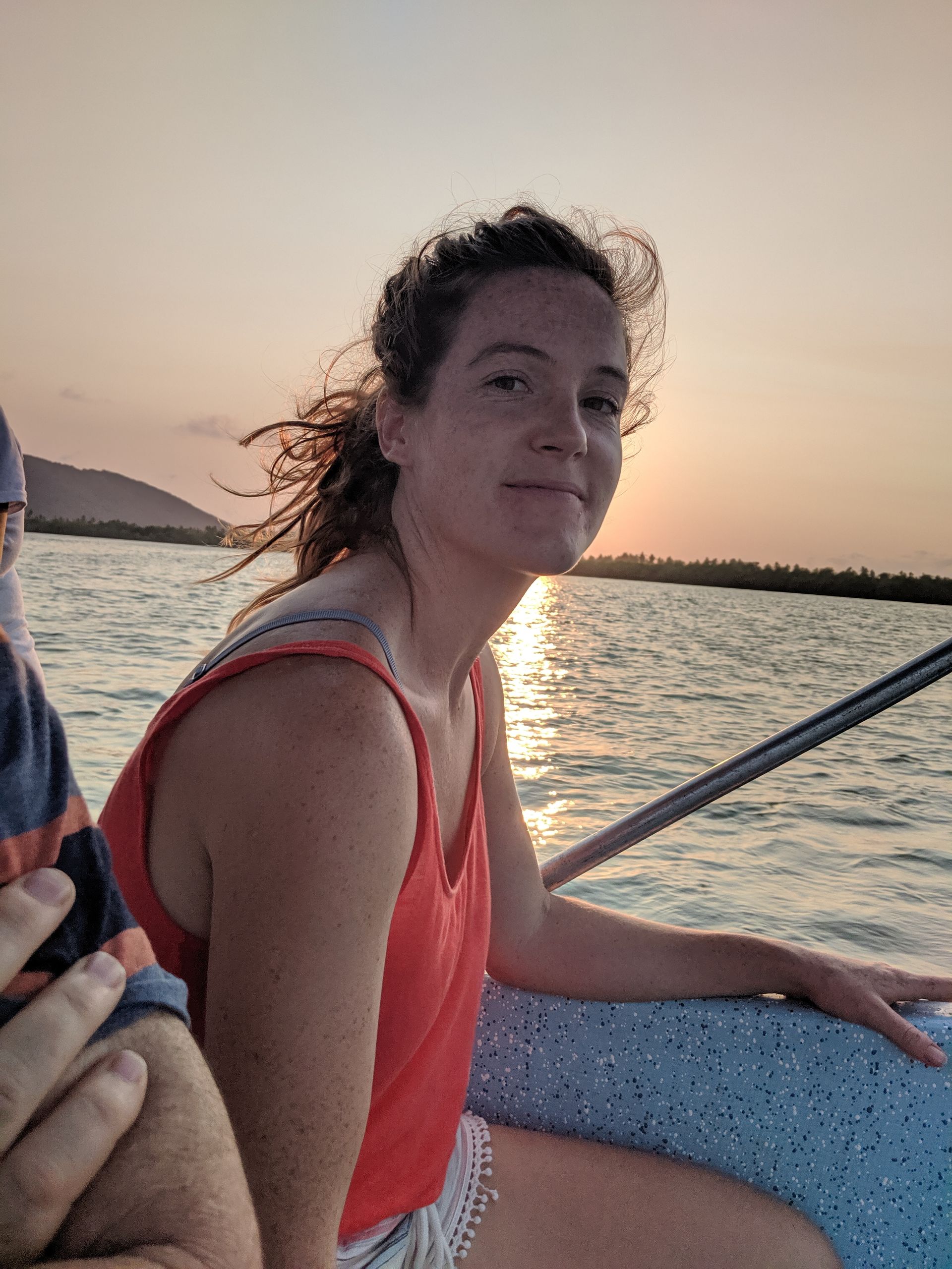 Woman in a coral top sitting on a boat at sunset, smiling by the water.