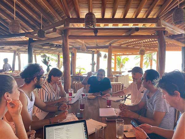 People gathered around a table in a rustic open-air café, talking and working on laptops.