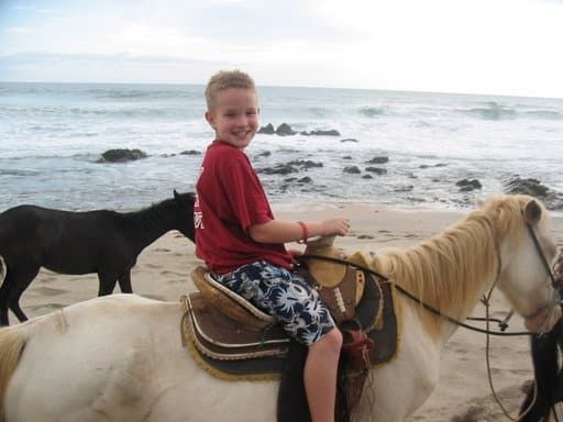 Child smiling on a white horse by the ocean, with a black horse nearby