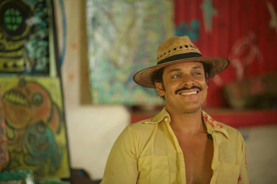 Smiling man in a straw hat and yellow shirt standing indoors with colorful paintings behind him
