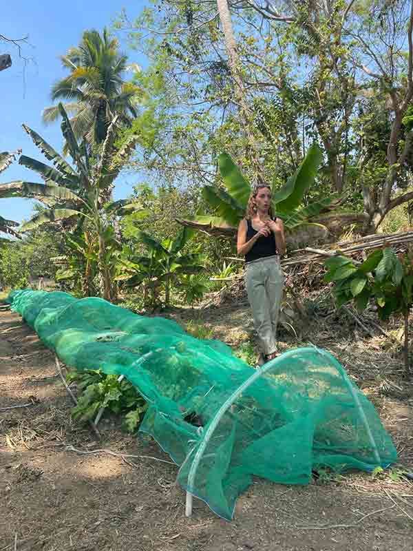 Person standing beside a green net tunnel in a tropical garden, holding their hands together.