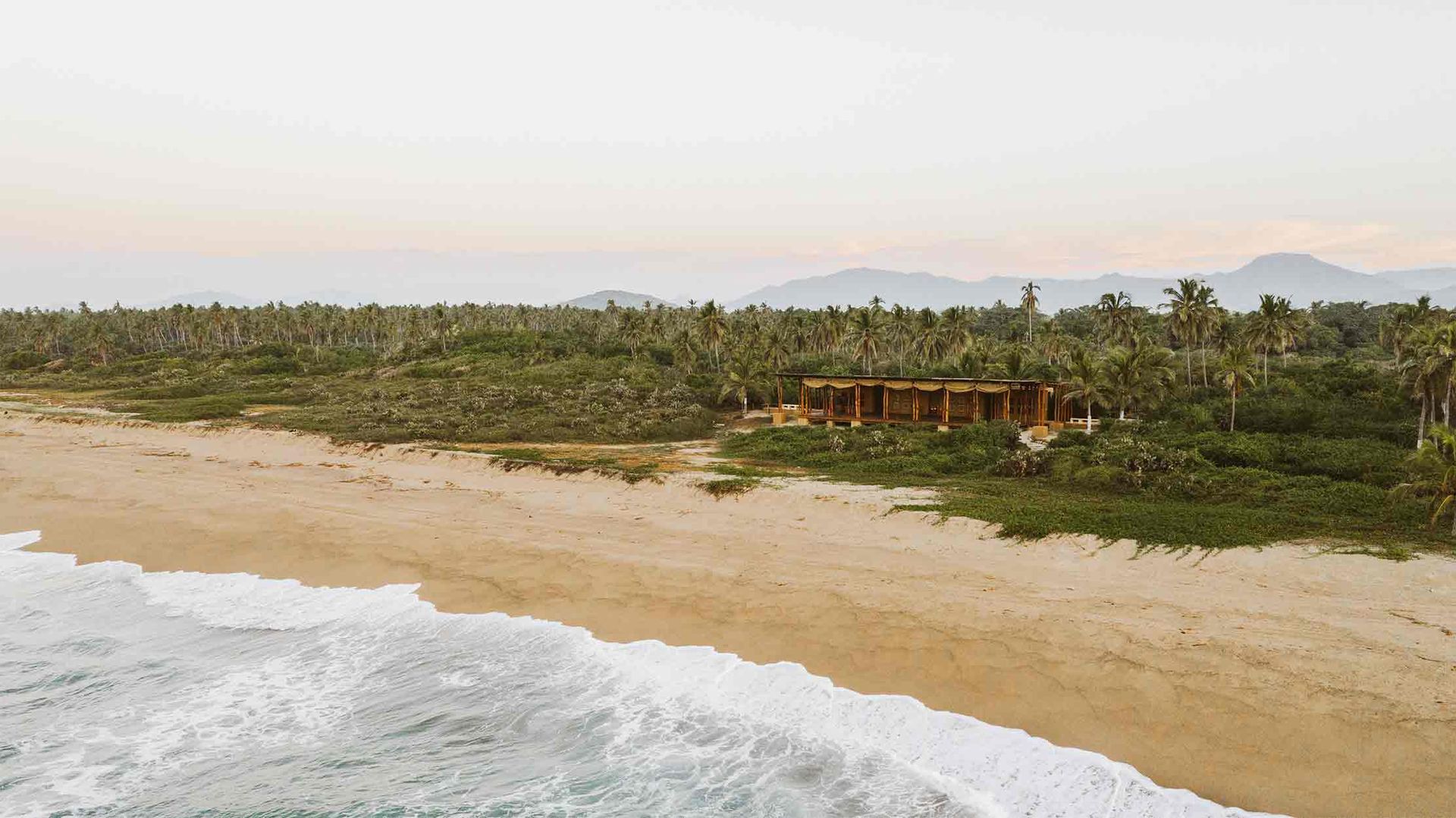 Sandy beach with waves, dense green shoreline, and small buildings near mountains under a pale sky