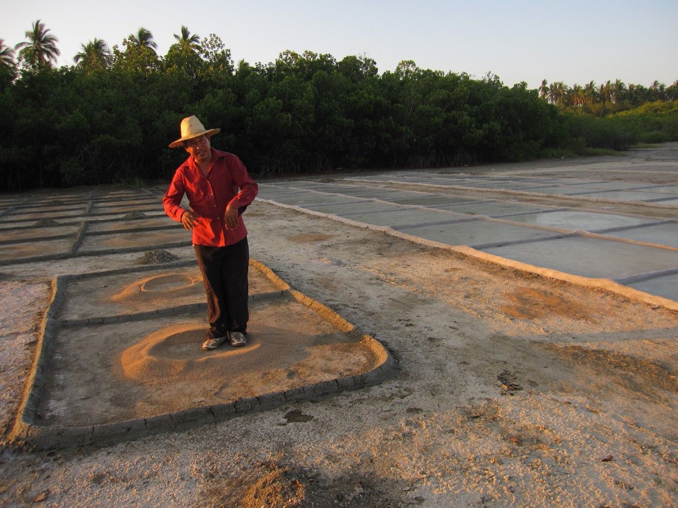 Person in a red shirt and straw hat standing on a gravel lot beside water and trees at sunset