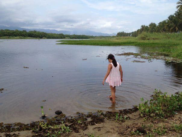 Person in a pink dress wades in a shallow lake beside a grassy shoreline under a cloudy sky.