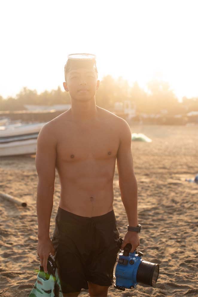 Shirtless person standing on a beach at sunset, holding camera gear and a green bottle