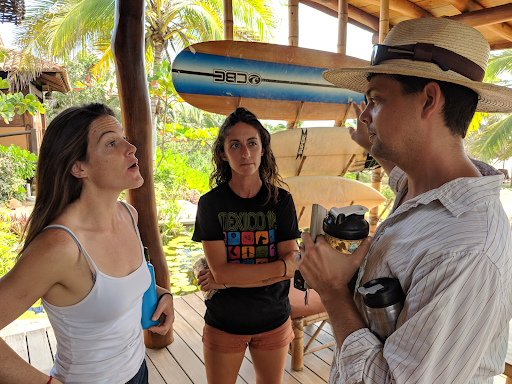 Three people talking in a beach hut, with a surfboard and palm trees in the background.