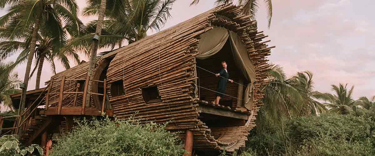 a bamboo treehouse in a lush tropical setting, with two people on the porch.