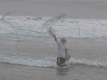 Person standing in shallow surf, raising a fishing net or line in gray water