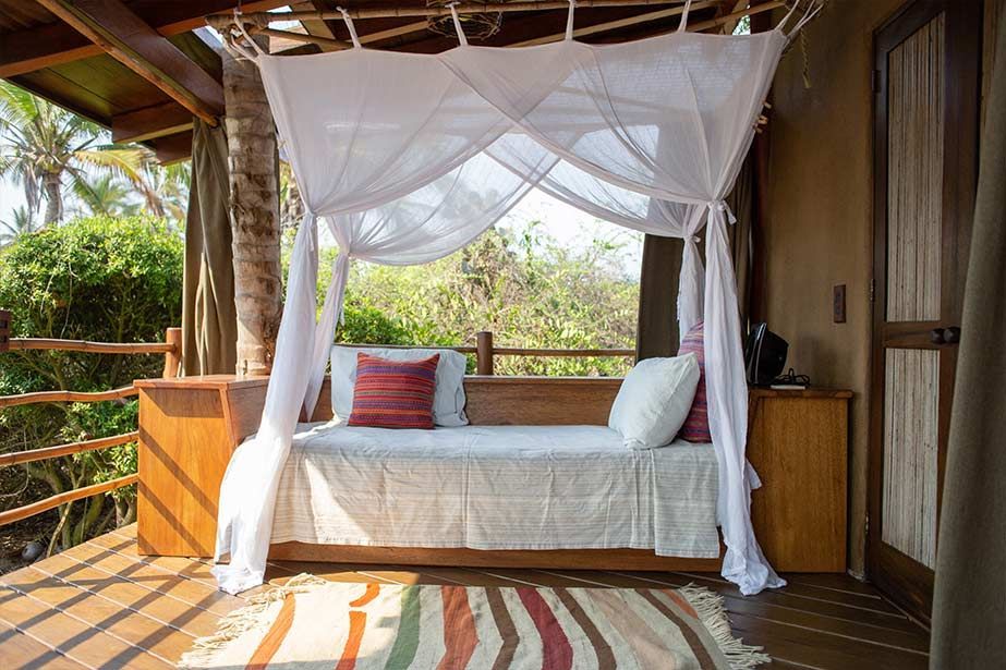 White canopy daybed on a wooden porch with colorful pillows and striped rug, overlooking greenery