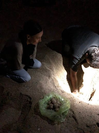 Two people crouch over a flashlight-lit spot in a dark room beside a green bag of small items.