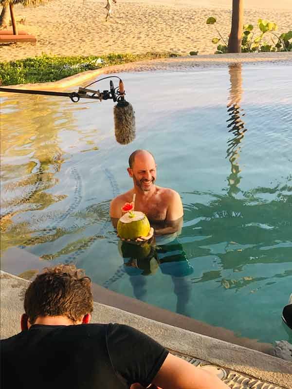 Man holding a yellow coconut drink in a pool, with another person leaning by the edge and beach nearby