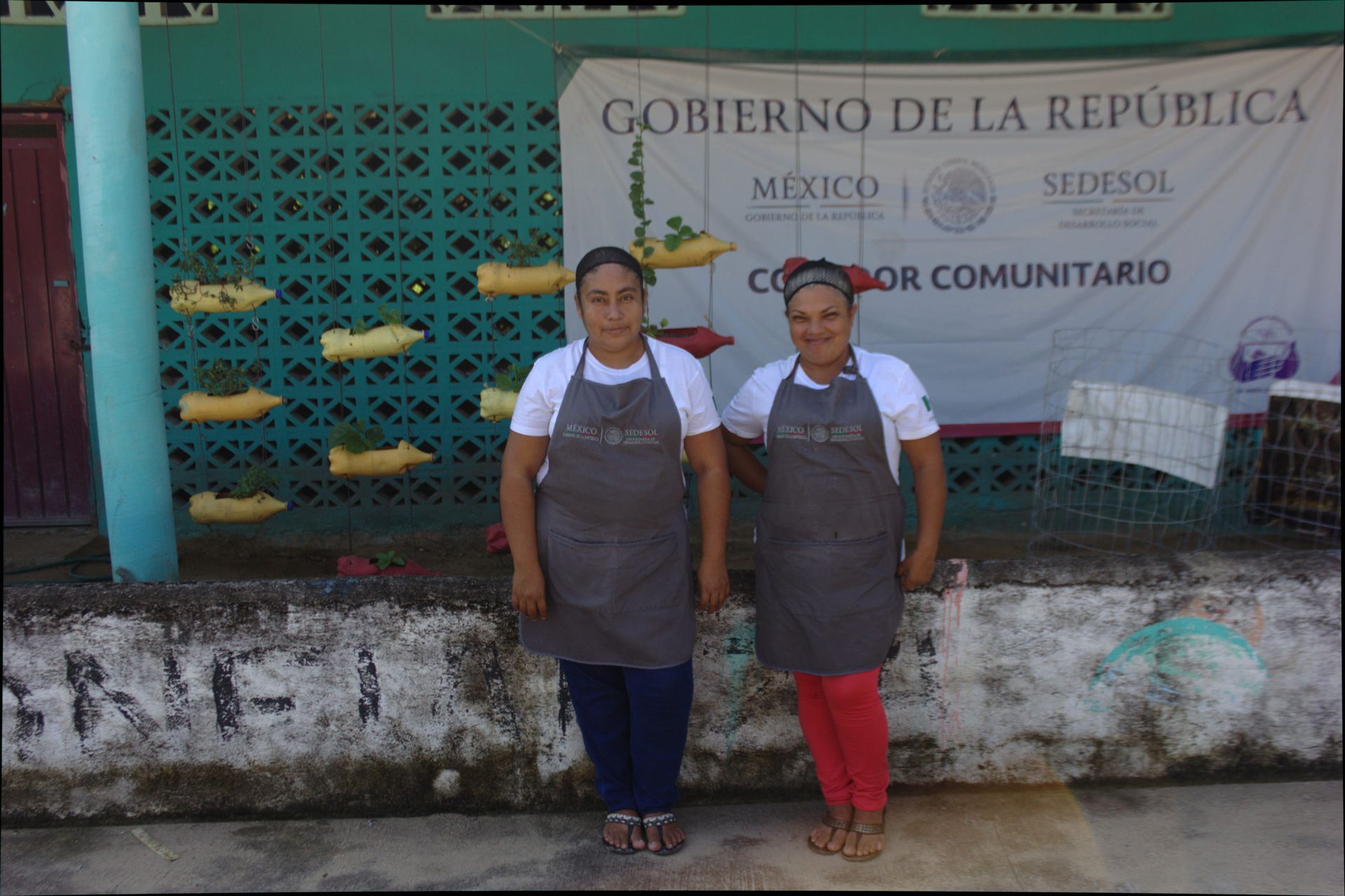Two women in aprons standing outside a community center with a Mexico government sign.
