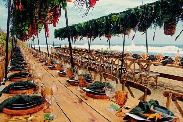 Beach dining setup with long tables, woven chairs, and green canopy drapes by the sea
