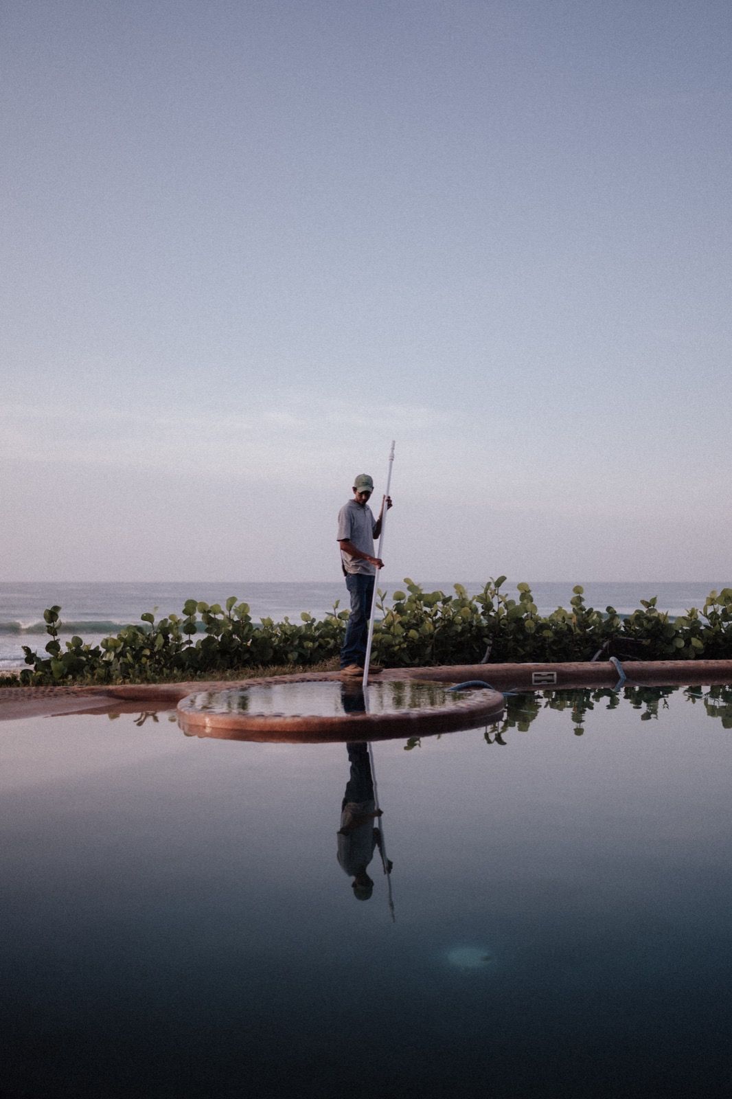 Person fishing beside a reflective infinity pool at dusk, with a calm ocean horizon beyond.