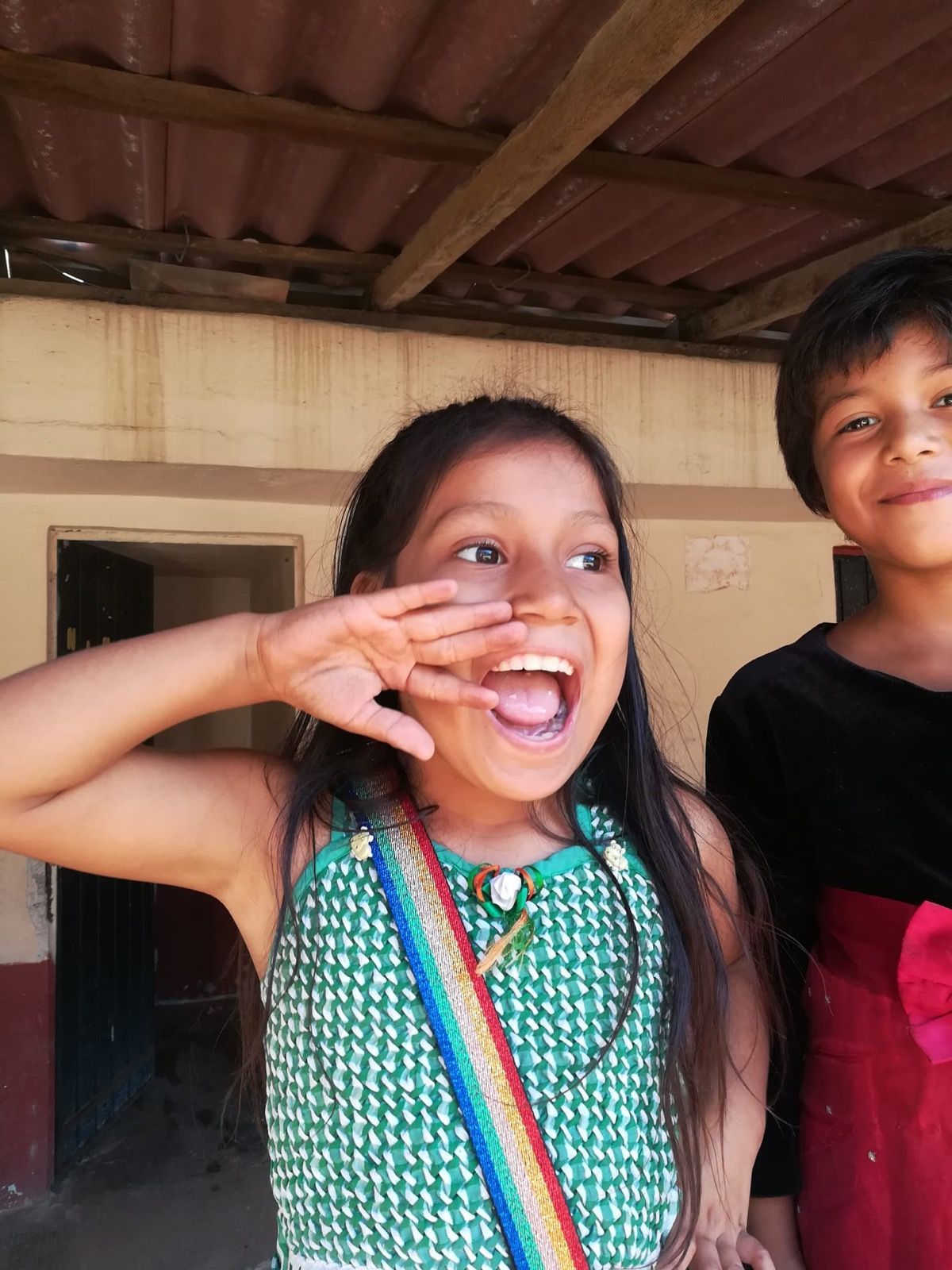 Two smiling children standing outdoors, one making a playful face with a colorful sash on her shirt