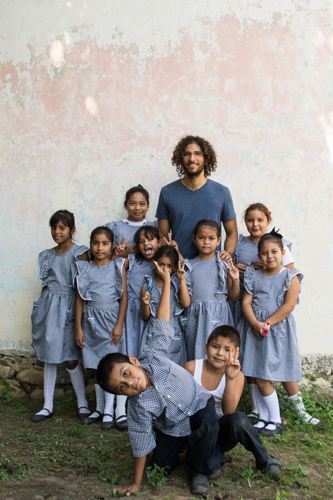 A group of children in blue matching outfits pose with an adult against a plain wall outdoors.