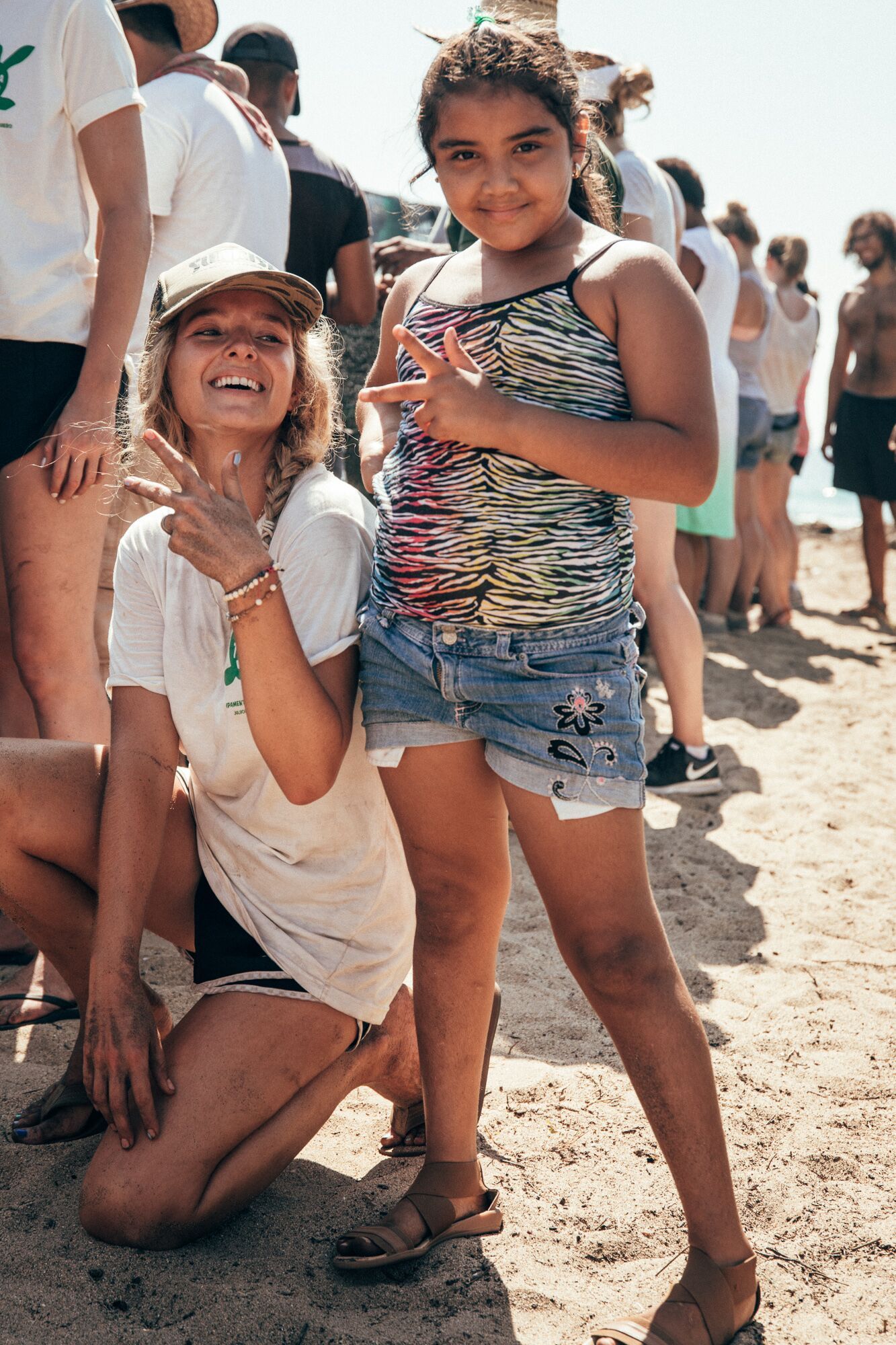 Two smiling people on a sunny beach, one kneeling and one standing, with other beachgoers behind them