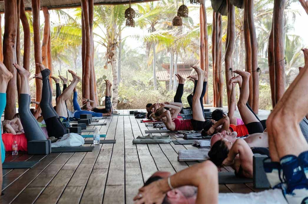 Group yoga class on mats in an open-air wooden pavilion, with people lying on their backs and legs raised.