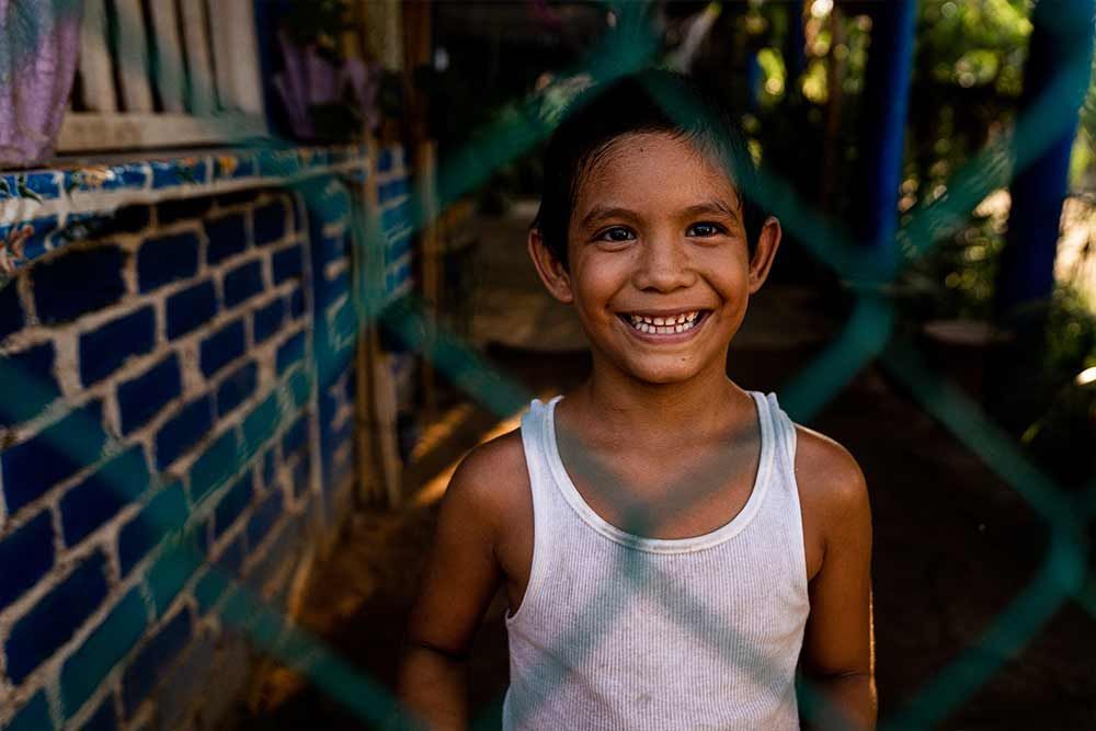 Smiling child in a white tank top standing by a blue brick wall behind a chain-link fence