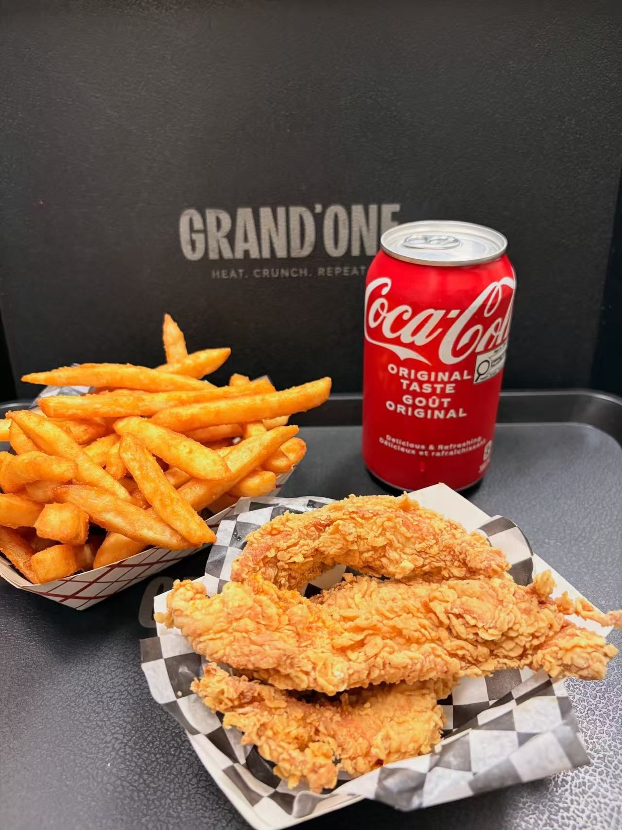 Fried chicken tenders, fries, and a Coca-Cola can on a checkered tray against a dark Grandon backdrop