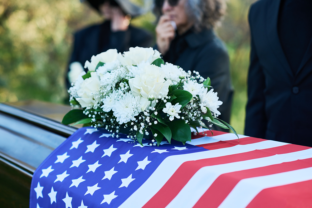 Casket draped with US flag, white flowers on top; people in black attire stand in the background.