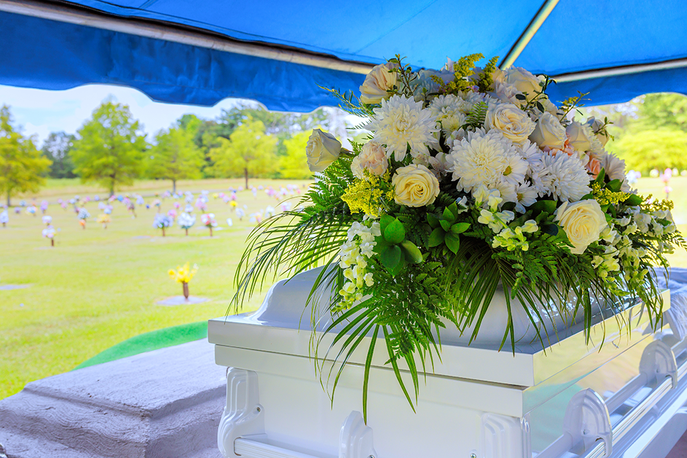 White casket with floral arrangement at a funeral, overlooking a cemetery with mourners.